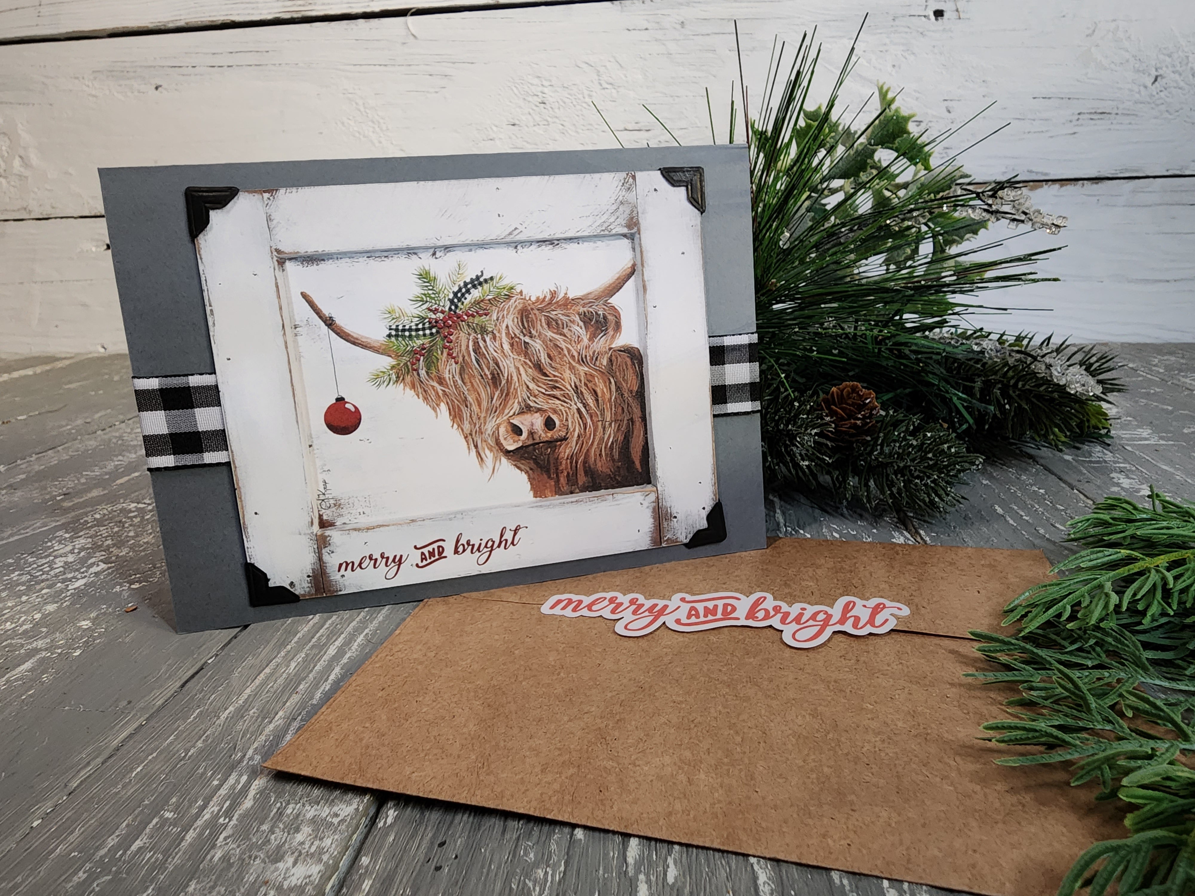 Single Handmade Christmas card with black and white buffalo plaid ribbon, Highland cow with Christmas evergreens, red berries, red bulb