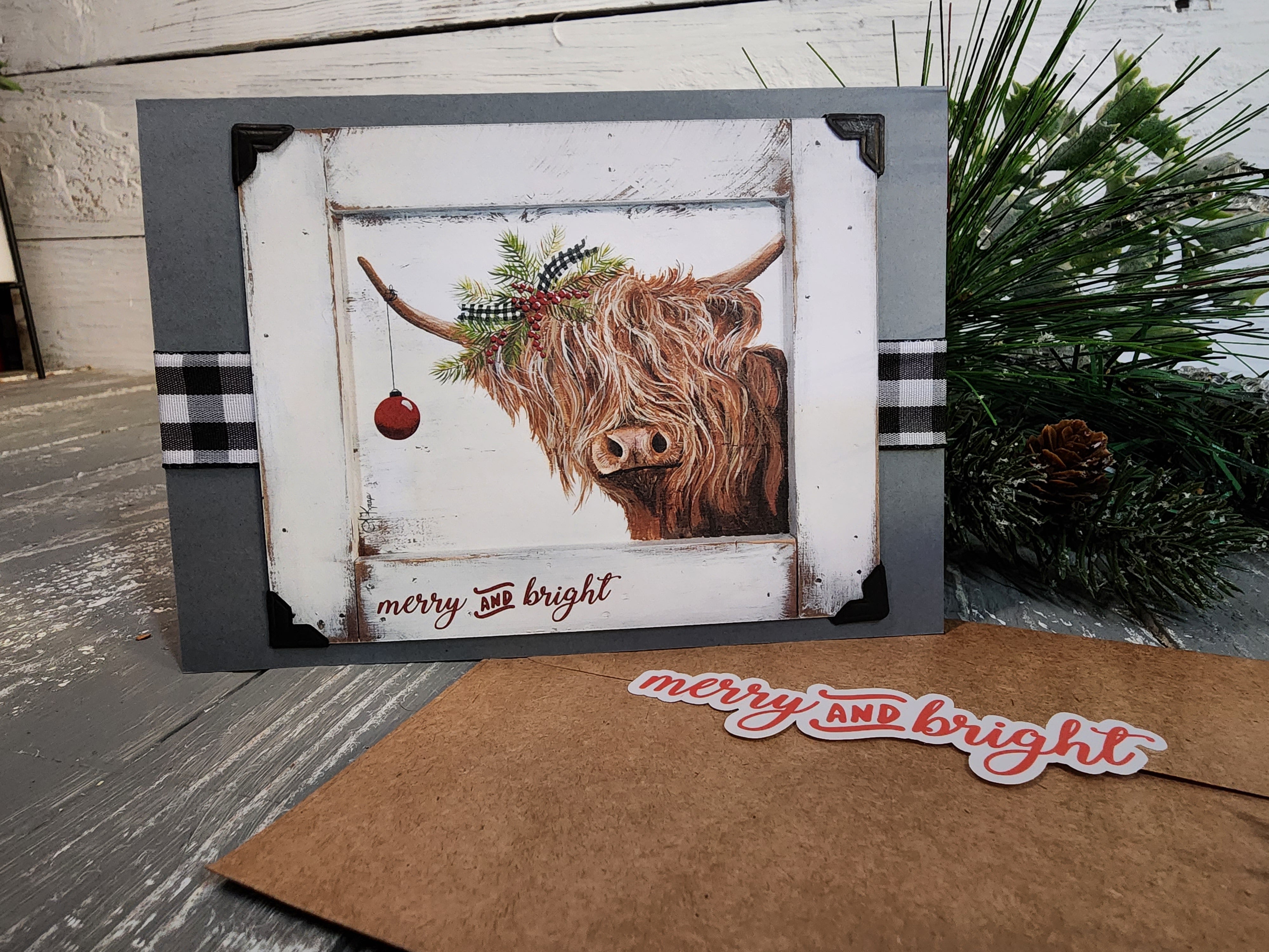 Single Handmade Christmas card with black and white buffalo plaid ribbon, Highland cow with Christmas evergreens, red berries, red bulb